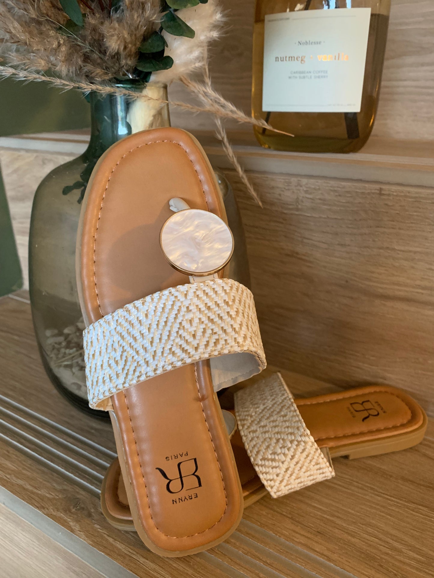 Zoom sur la sandale plate Luna Chic avec anneau nacré et bande tissée beige, posée élégamment sur fond bois clair avec vase de décoration naturelle