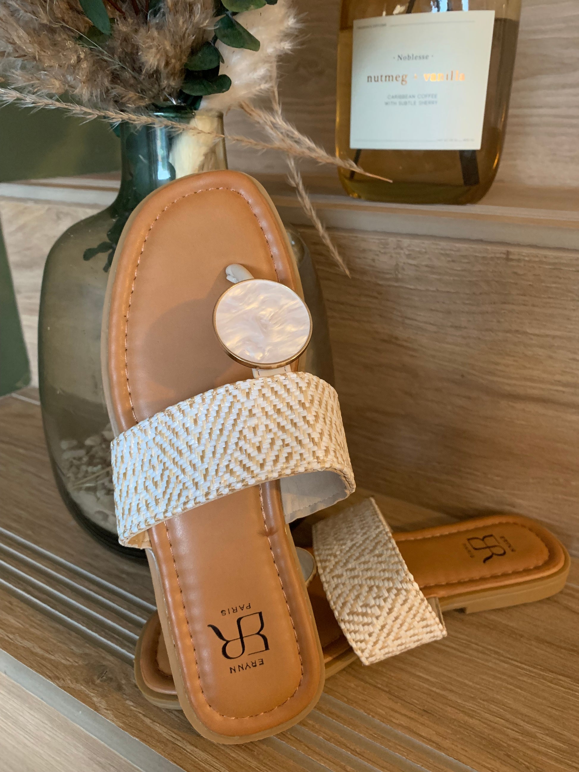 Zoom sur la sandale plate Luna Chic avec anneau nacré et bande tissée beige, posée élégamment sur fond bois clair avec vase de décoration naturelle